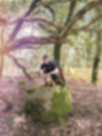 Will Thompson and his dog Dizzy in a forest on Dartmoor. Will is smiling at the camera and perched on a rock.