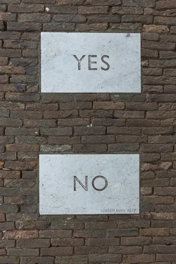 White plaques on a brick wall with "YES" and "NO" engraved. Below, inscription reads "Willem Buys 1977". Contrast creates a contemplative mood.