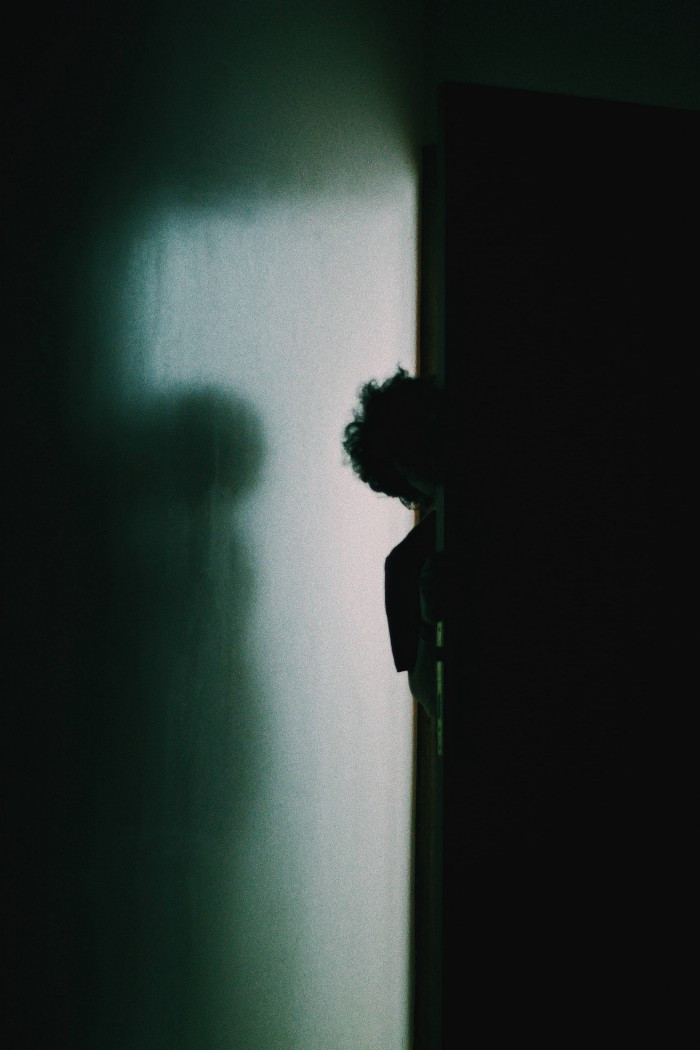 A boy in a barely lit hallway peaking around the corner into a very dark room.