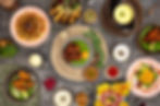 Overhead view of a colorful Indian feast: multiple plated dishes, sauces, spices, and herbs on a gray tablecloth, no people