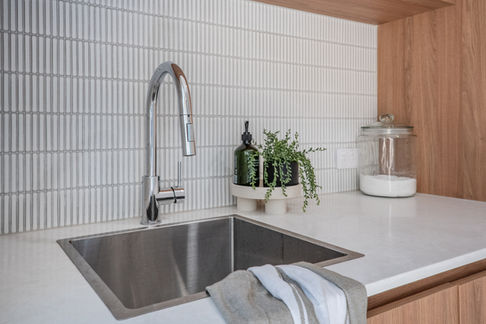 Close-up of a modern laundry room sink area in Perth, with wooden cabinetry, a white benchtop, a stainless steel sink, and a grey tiled splashback