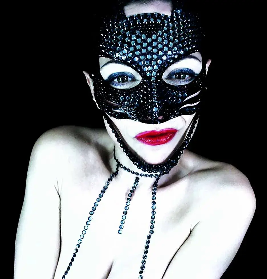 Una donna con una maschera nera tempestata di gioielli e labbra di un rosso acceso posa contro uno sfondo scuro, emanando una presenza misteriosa e suggestiva. Immagine molto contrastata con riverberi blu. Fotografia artistica e maschera sono opere di Antonella Sportelli