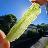 Hand holding a translucent green Moldavite crystal against a bright sunny sky.