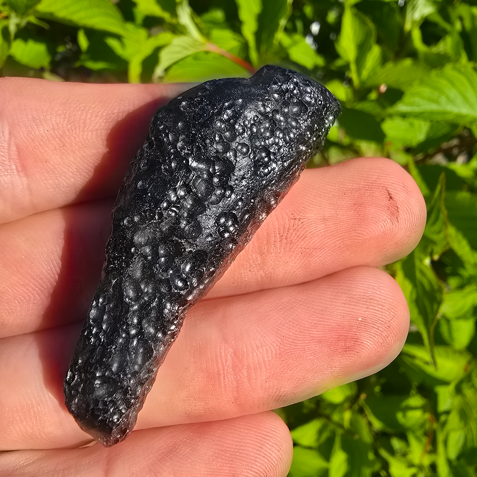 Thumbnail: Hand holding a piece of dark, textured Libyan Desert Glass