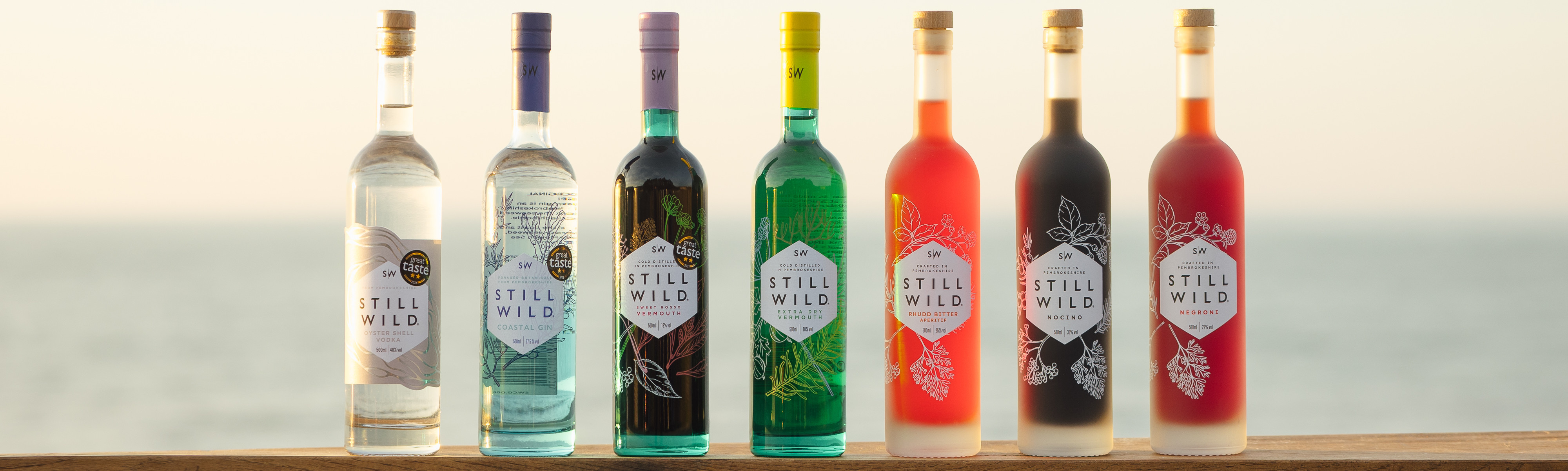 Seven Still Wild spirit bottles on a wooden ledge by the sea, with colorful labels and soft sunset light.