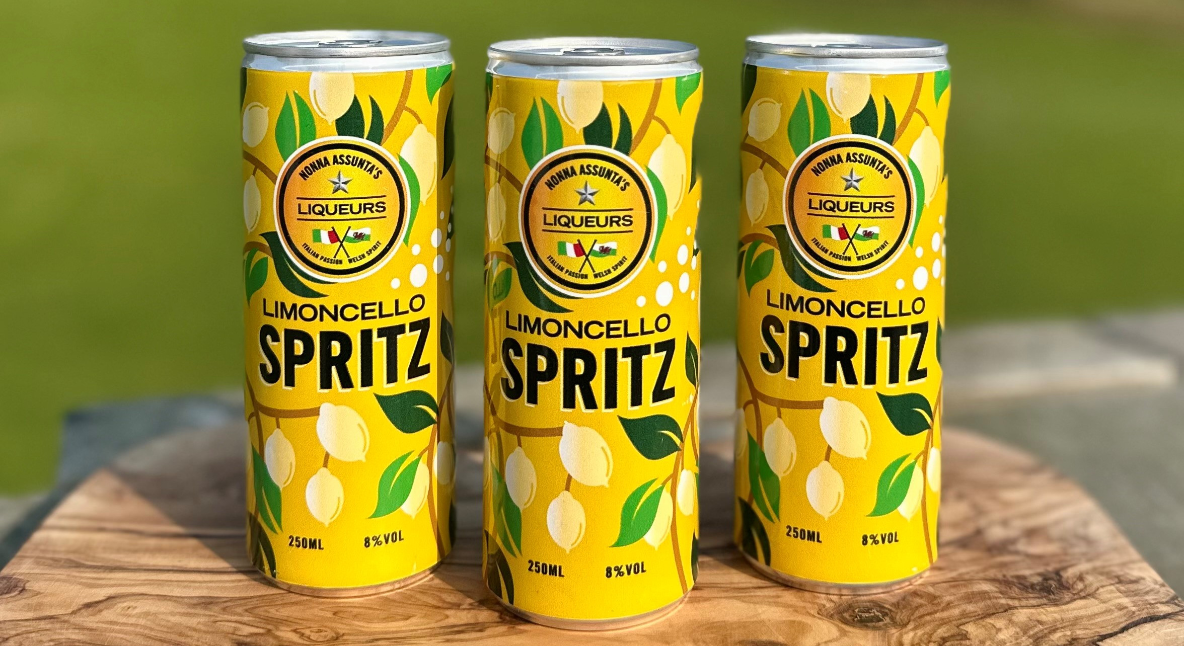Three yellow Limoncello Spritz cans on a wooden table outdoors, with a blurred green park behind them.