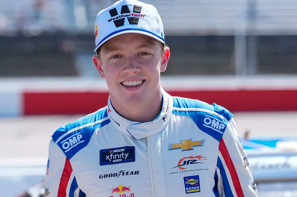 Race car driver in white and blue suit with sponsor logos smiles, wearing a cap. Background shows blurred racetrack and stands.