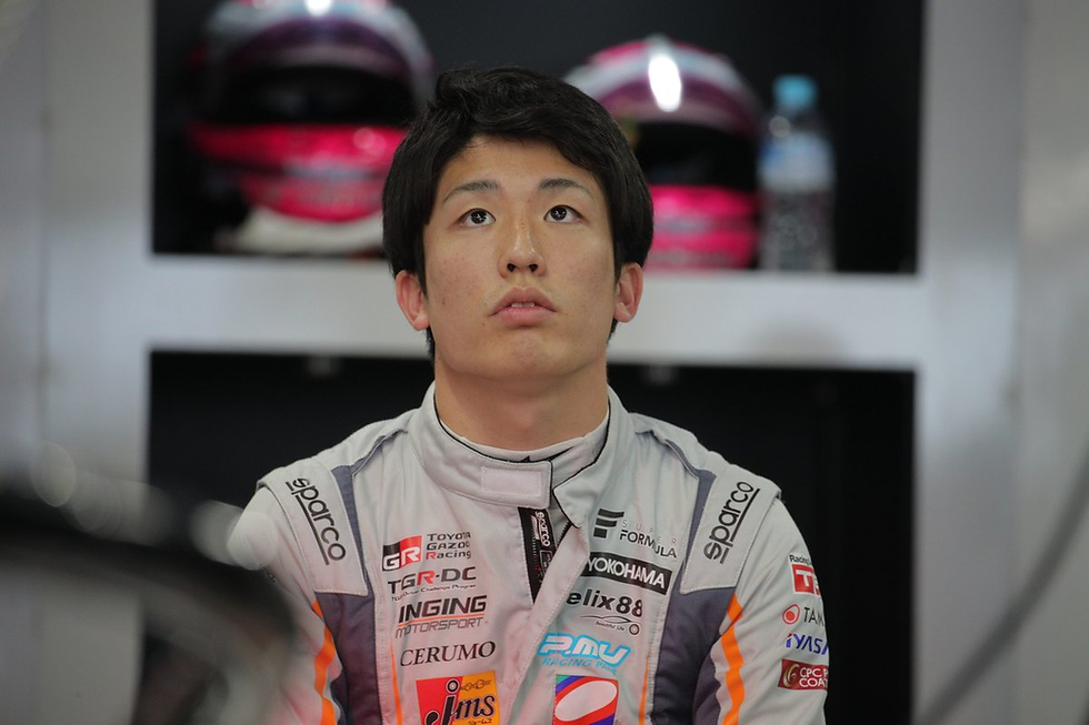 Race car driver in a branded suit looks up with a focused expression. Background shows helmets and a water bottle on shelves.