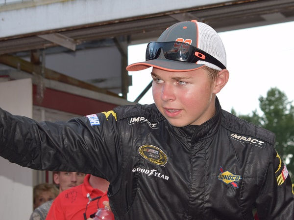 Young race car driver in black suit and cap gestures confidently, surrounded by people and greenery. Logos: Sunoco, Goodyear, Impact.