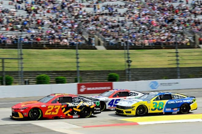 Race cars numbered 23, 45, and 38 speed around a track. The vibrant designs include red, blue, and yellow. A crowd fills the stands.