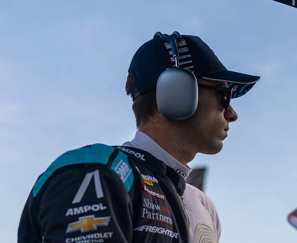 Person in racing gear with sponsor logos, wearing headphones and sunglasses, stands against a clear blue sky, looking focused.