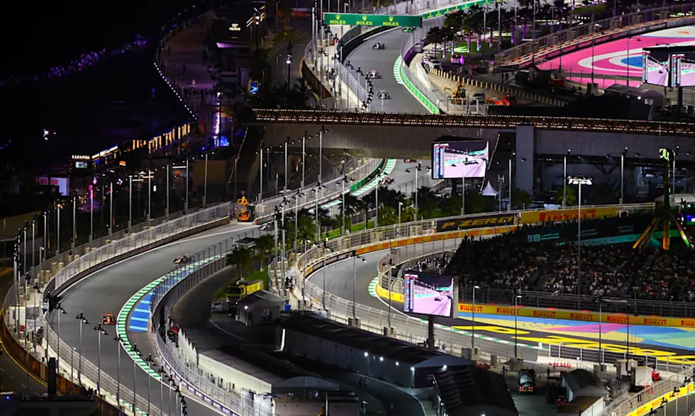 Nighttime car race on a lit track with colorful markings, surrounded by lights and spectators. "Rolex" signs visible above the track.