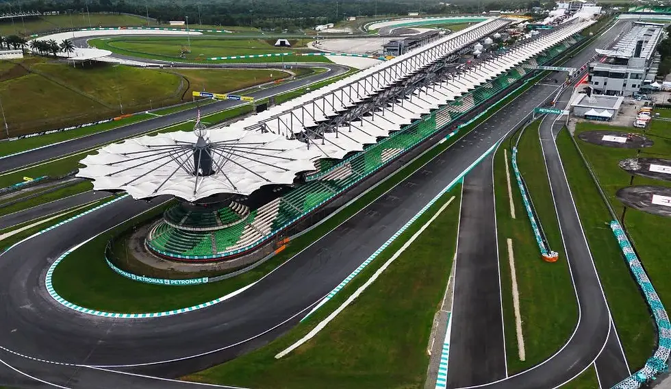 Aerial view of an empty racetrack with green and white stands, a distinctive canopy structure, grassy surroundings, and "Petronas" signage.