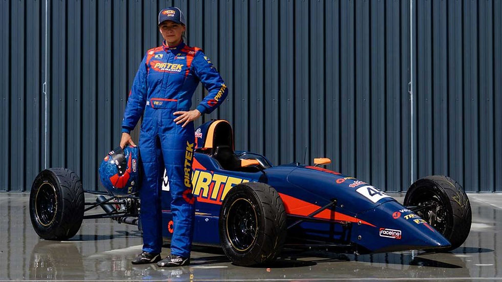 Racer in blue suit stands confidently beside a blue and orange race car labeled "PIRTEK", against a gray corrugated metal backdrop.