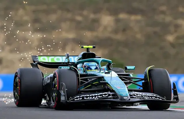 Formula 1 car racing on track, sparks flying from the rear. The vehicle is green with logos, set against a blurred background, emphasizing speed.