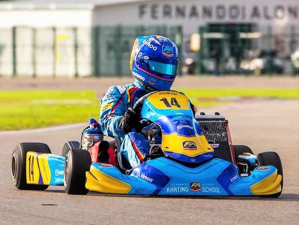 Person in blue racing gear drives a go-kart on a track. "KARTING SCHOOL" text visible. Vibrant yellow and blue colors, sunny day.