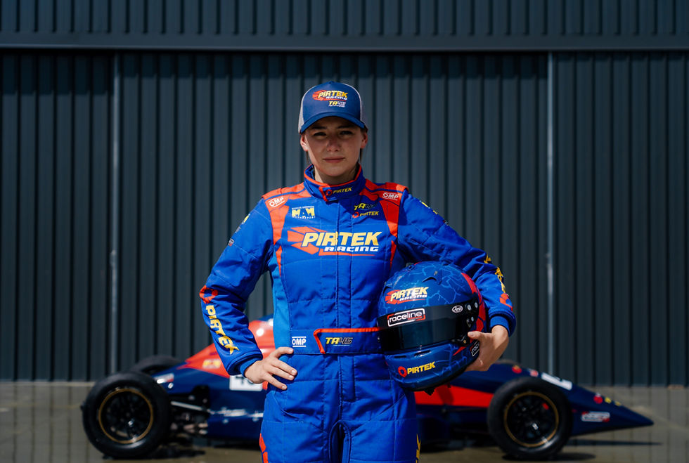 Racer in blue and red Pirtek suit holds helmet next to formula car against a corrugated metal backdrop. Determined expression.
