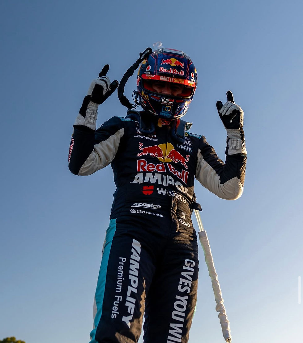 Race car driver in a Red Bull suit poses confidently with raised fingers against a clear blue sky, exuding triumph and excitement.