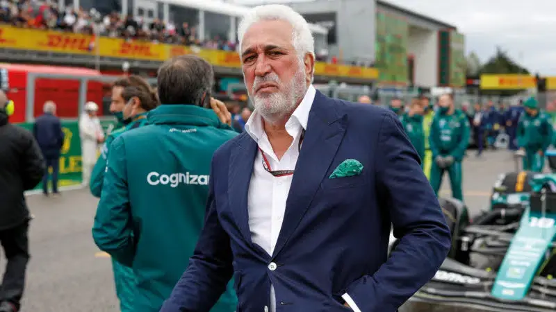 Man in a navy suit stands confidently on a racetrack, surrounded by people in green uniforms. A DHL sign is visible in the background.