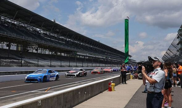 Race cars on a track, led by a blue car, under a cloudy sky. Spectators and a photographer are on the sidelines. Green flag visible.