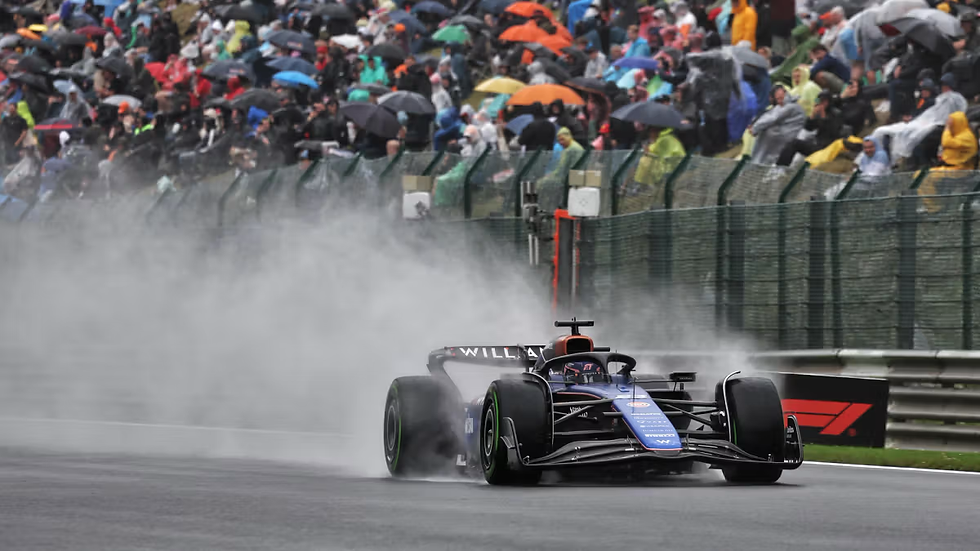 Race car on wet track sprays water; crowded stands with colorful umbrellas in background. Car labeled "WILLIAMS," rainy atmosphere.