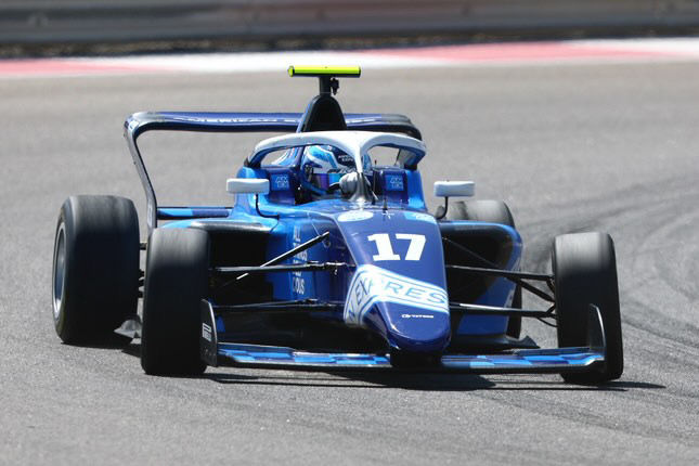 Blue race car number 17 speeds on a track with visible tire marks. Driver in helmet is focused. Bright, sunny day enhances the action.