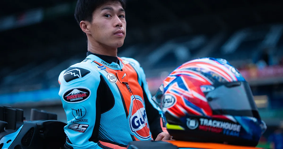 Motorcyclist in blue and orange suit looks back, standing by a colorful helmet in a racetrack setting. Overcast mood; helmet text: Trackhouse.