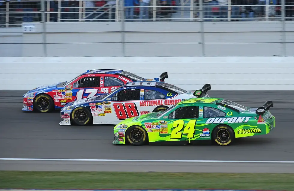 Three colorful race cars, numbers 24, 88, and 17, speed side by side on a track with spectators in the background.