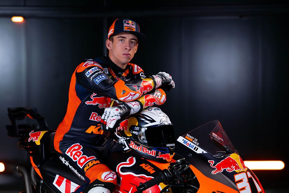 Motorcyclist with orange and black gear leans on a racing bike in a dimly lit room. Visible logos: Red Bull, KTM. Determined expression.