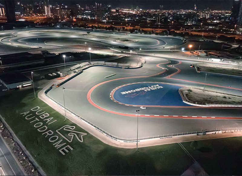 Aerial view of Dubai Autodrome at night, lit track with curves, red and blue lines, city skyline in background, logo visible on ground.