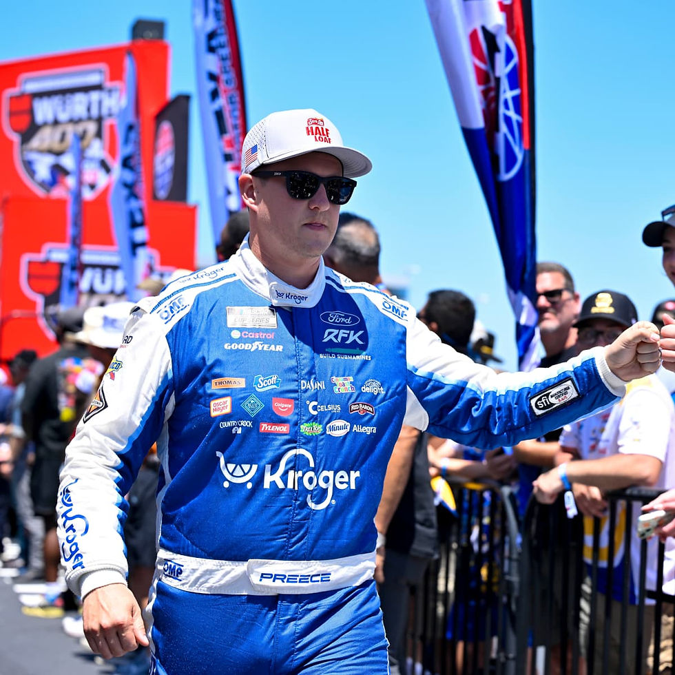Race car driver in blue Kroger suit fist-bumps a fan. Crowd and red/blue banners in the background. Bright, sunny day.