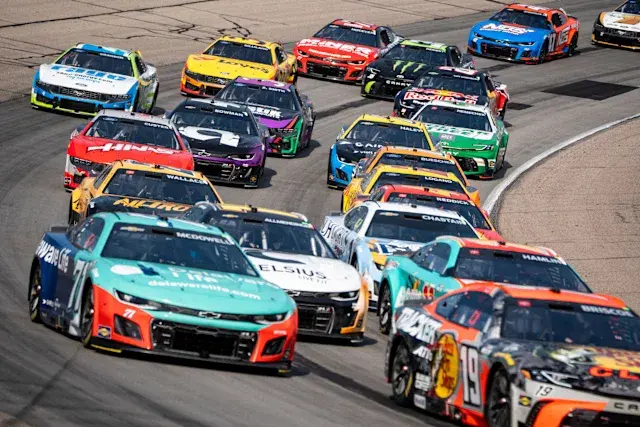 Race cars in vibrant colors speed around a track, tightly packed and competing. The asphalt and white lines highlight the intense action.