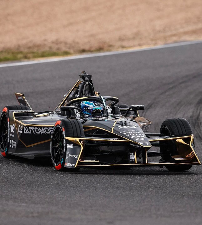 Black and gold race car with "DS Automobiles" on the side is speeding on a racetrack. Jess in blue helmet, cloudy background.