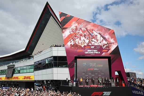 Large crowd at the British Grand Prix podium ceremony. A vibrant red and black backdrop features racers, under a partly cloudy sky.