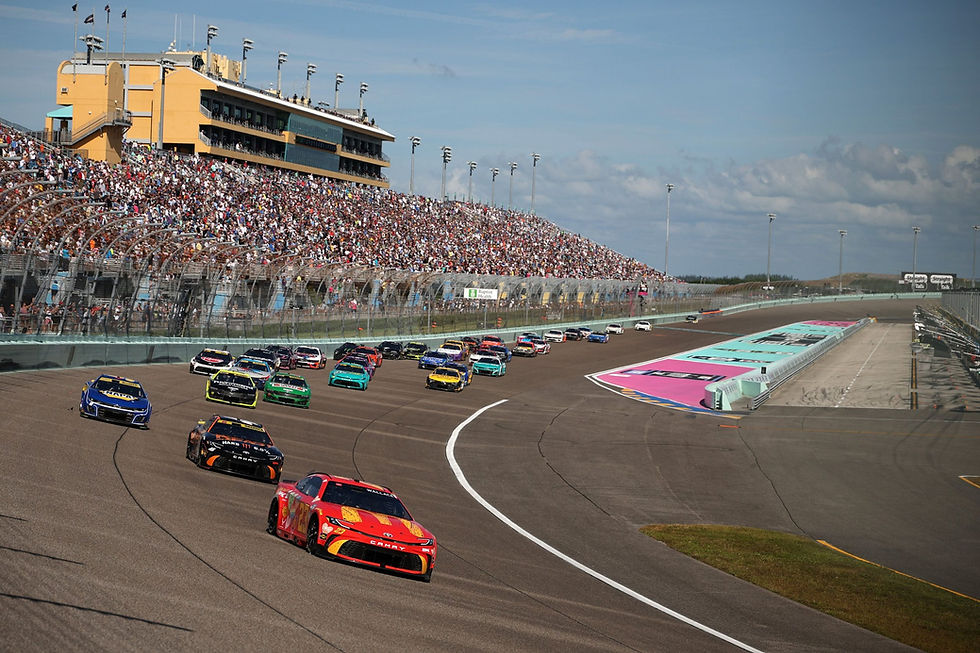 Race cars speed on a track, leading a dynamic turn. A packed grandstand and large building are visible; the scene is vibrant and energetic.