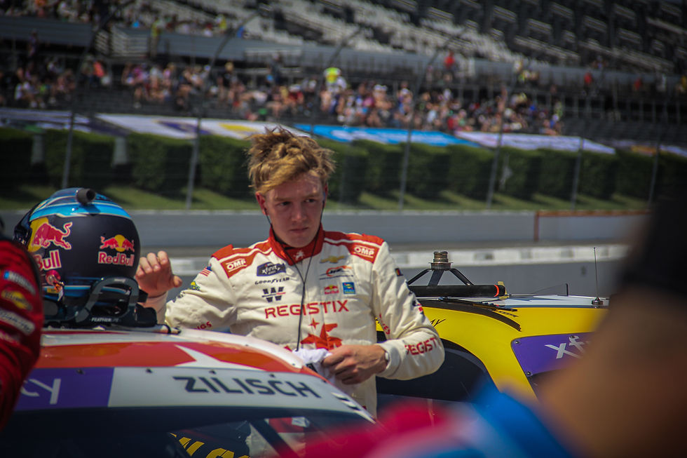 Race car driver in white suit adjusts helmet beside yellow car on a track. Stands with crowd in background, focused expression.
