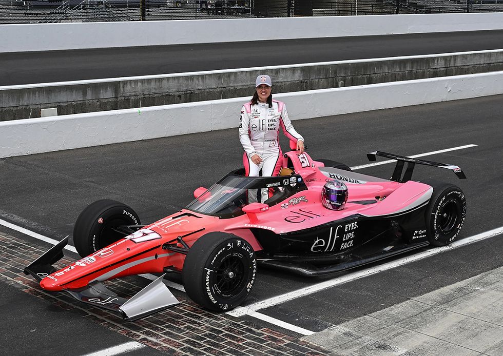 Racecar driver in white and pink suit poses with pink-black car on track. "elf EYES. LIPS. FACE." on car. Helmet on car.