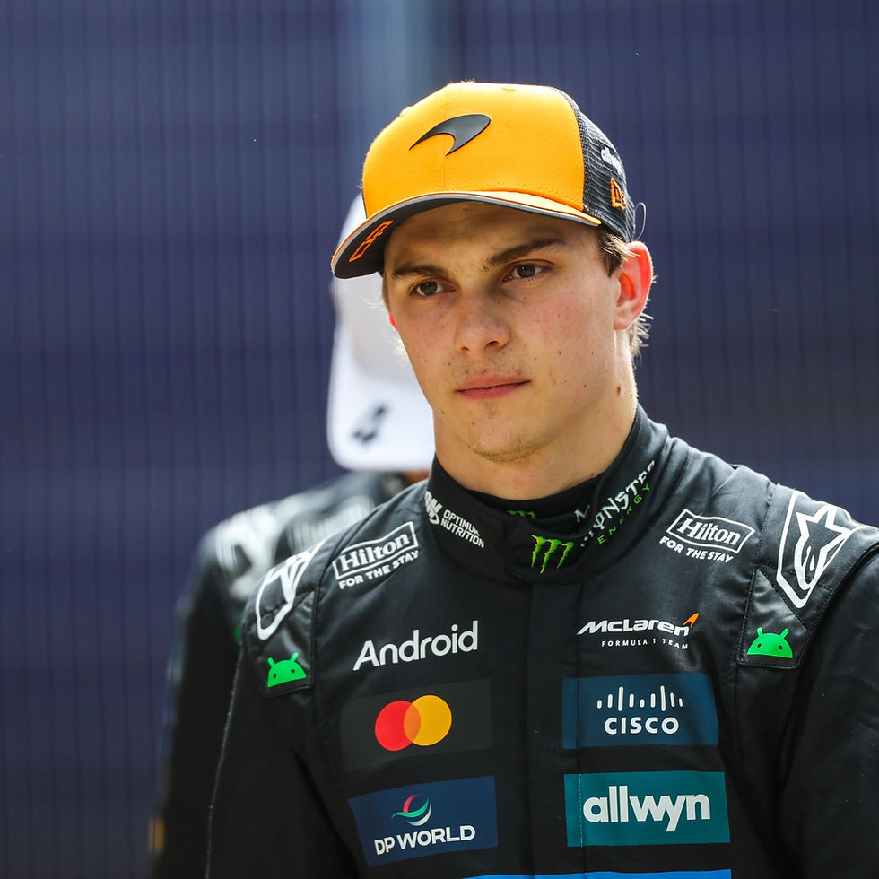 Race car driver in a black and orange cap and suit with sponsor logos like Android, McLaren, and Cisco. Serious expression, blurred background.