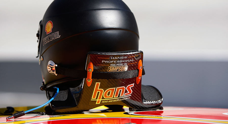 Black helmet on a colorful car with "Hans" and "Simpson" text, showing safety straps. Pennzoil and Shell logos visible, sleek design.