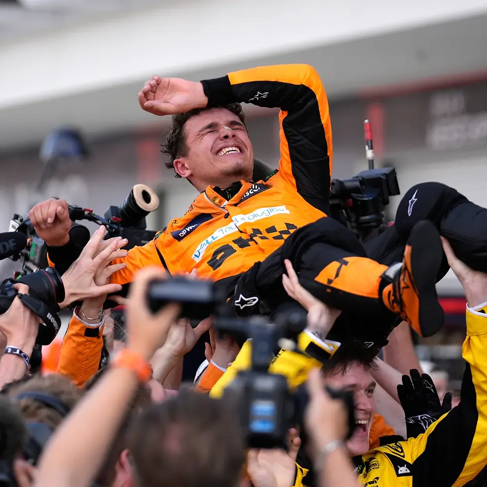Race car driver in orange suit is lifted by a jubilant crowd, smiling and celebrating. Cameras capture the moment, creating a lively scene.