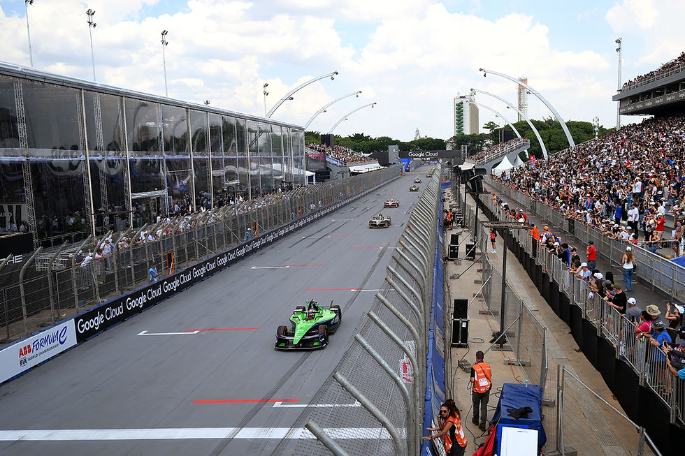 Race cars speed on a track with a green car in front. Crowd watches from bleachers. "Google Cloud" ads visible. Energetic atmosphere.