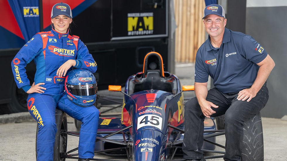 Young woman in blue racing suit and man in blue cap and shirt smile beside a race car. Logo "PIRTEK" visible. Background: racing garage.
