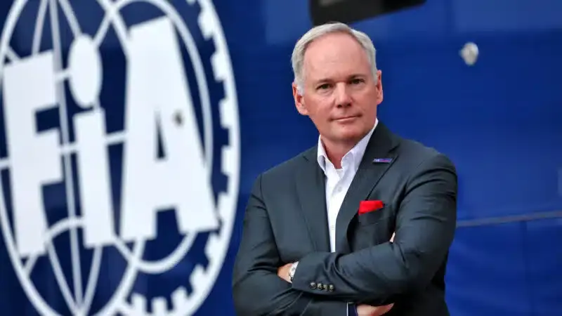 Man in a dark suit stands with arms crossed. Background features large FIA logo on blue. Mood is professional and confident.