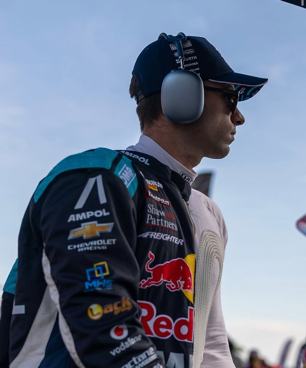 Race car driver in branded suit and headphones stands against a clear sky, with logos like Chevrolet and Red Bull visible, conveying focus.
