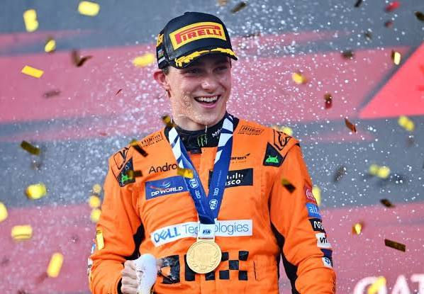 Racing driver in orange suit and Pirelli cap smiles with gold medal, surrounded by confetti on a podium. Energetic celebration scene.
