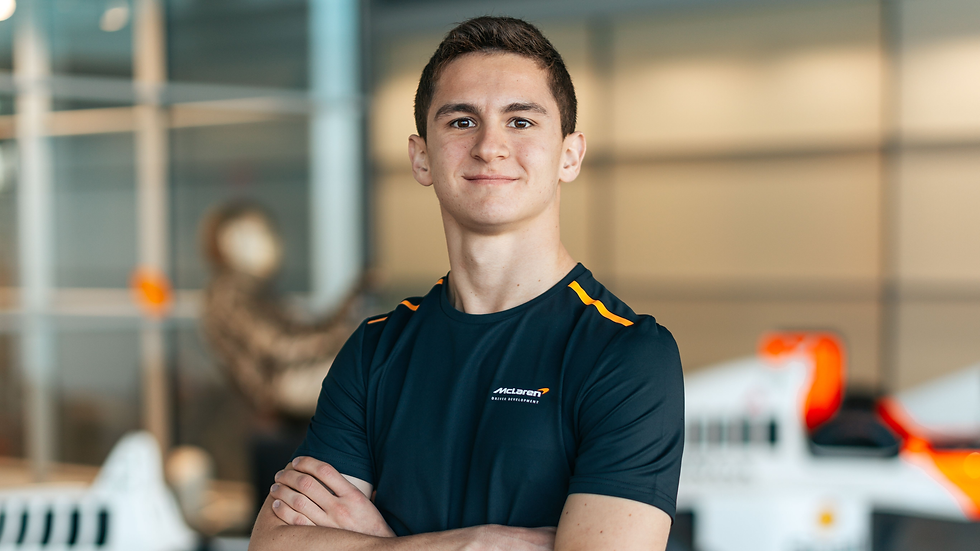 Young man with crossed arms in a McLaren shirt, smiling in a modern setting with blurred racing car elements in the background.