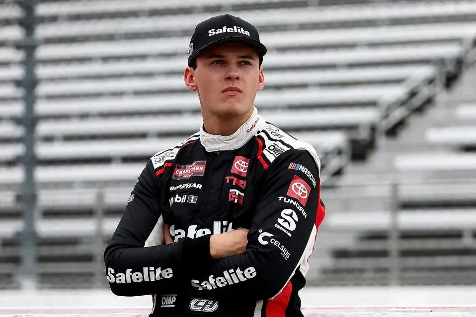 Race car driver in a black Safelite suit and cap, arms folded, stands confidently at a racetrack. Empty bleachers in background.