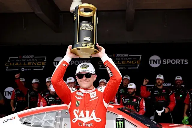 Race car driver in red suit holds trophy overhead, smiling. Team celebrates behind. Car and sponsor logos visible. Jubilant mood.