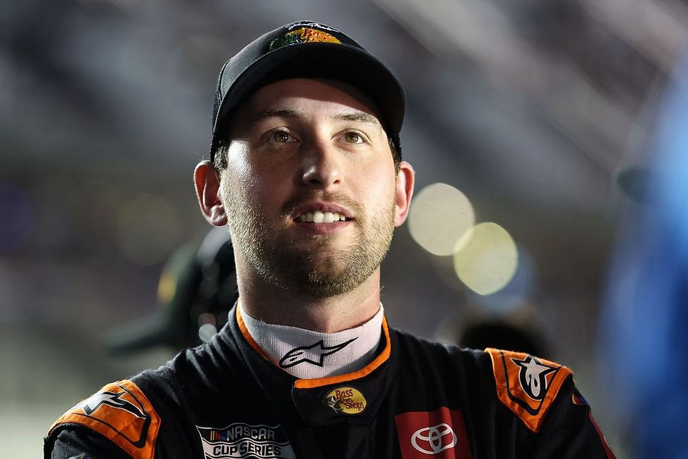 A race car driver in a black and orange suit smiles under bright lights. The NASCAR Cup Series logo is visible on the suit.