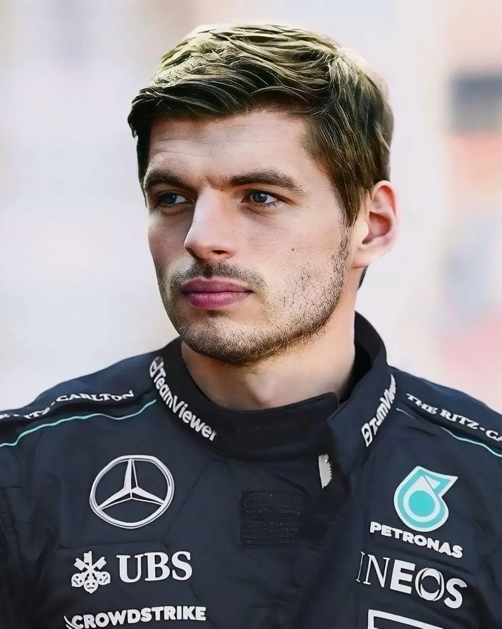 Race car driver in a black suit with logos, looking serious. Background is a blurred outdoor scene. Logos include Mercedes, UBS, Petronas, INEOS.